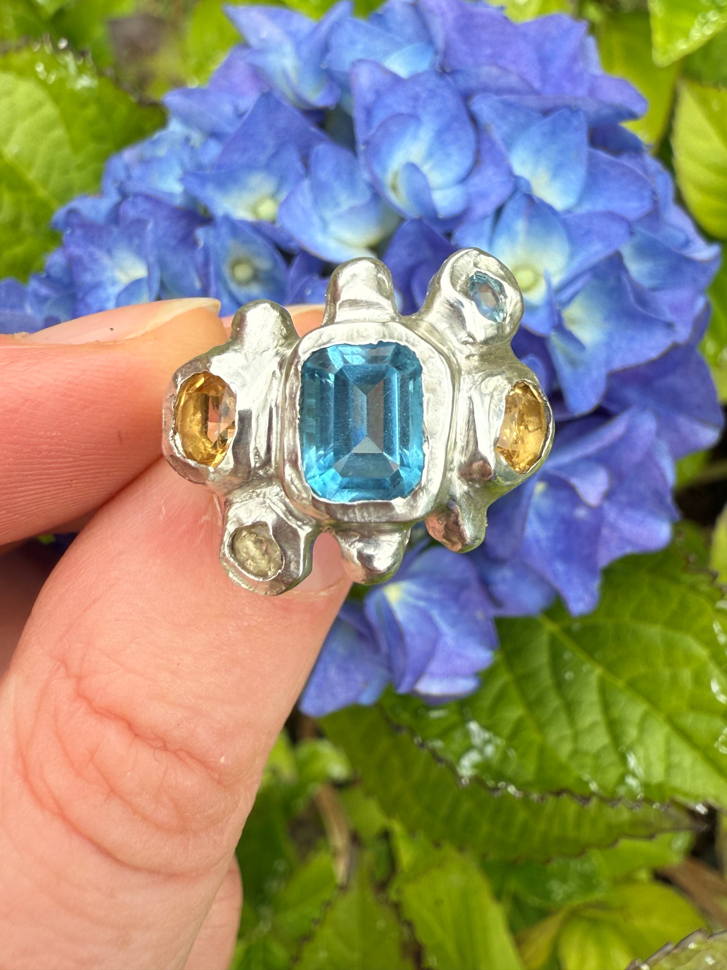Silver bohemian ring with blue and yellow gemstones held against a background of blue flowers and green leaves