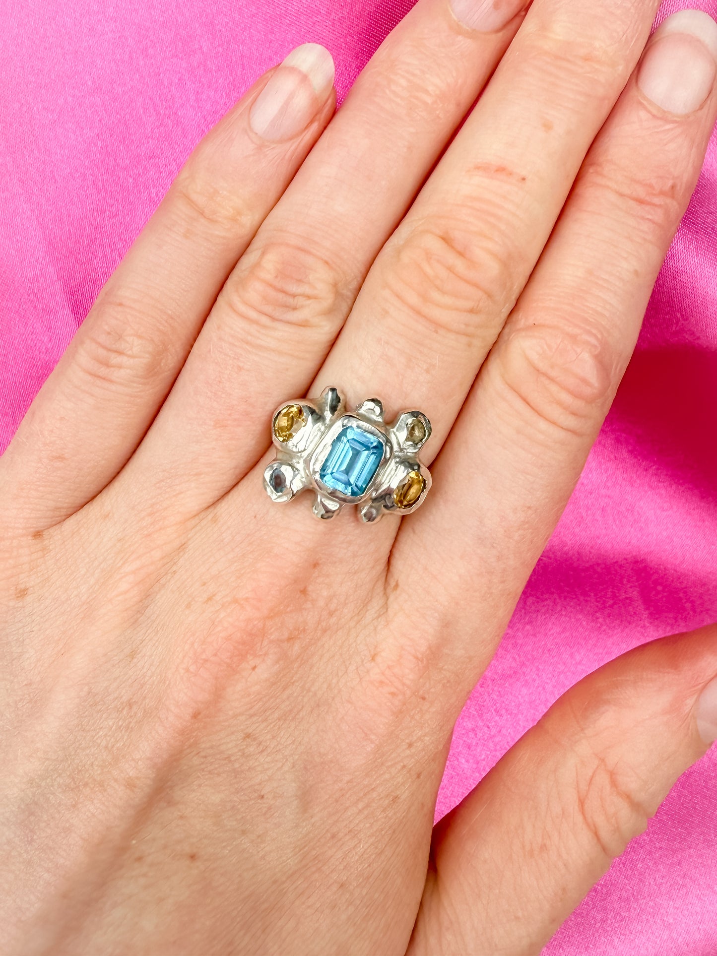 Silver Ring with blue and yellow gemstones on a hand against a pink background