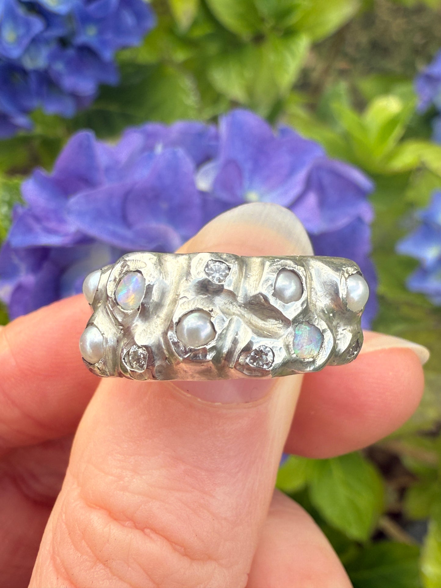 Silver ring with pearl, opal and moissanite stones held by a hand against a background of blue flowers.