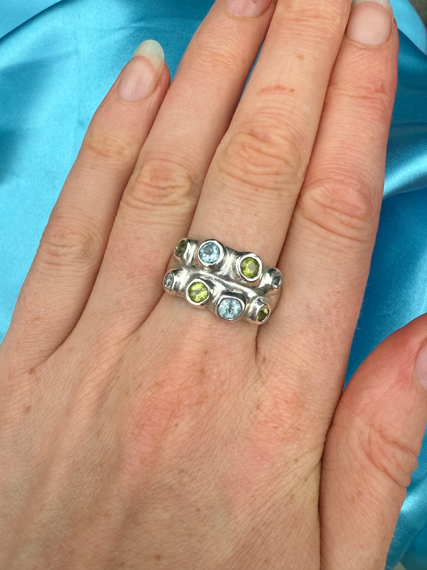 Silver bohemian ring with blue and green gemstones on a hand against a blue satin background