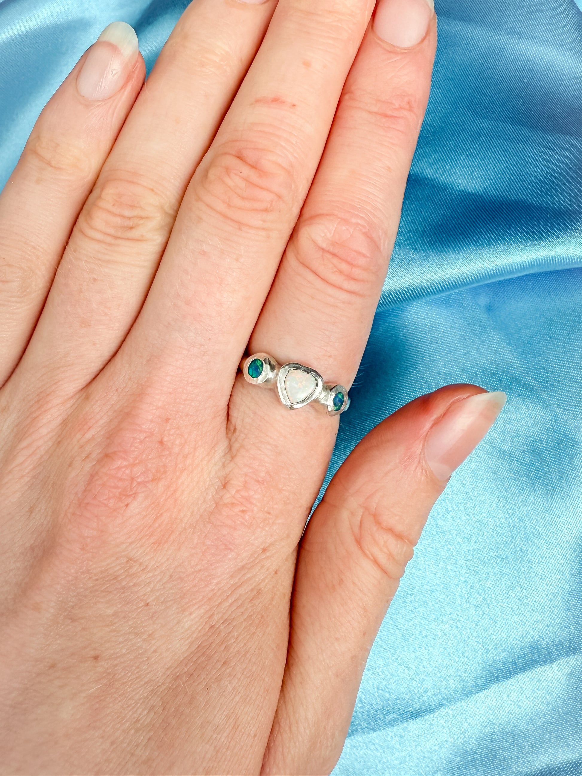 Hand wearing a silver ring with heart design and blue gemstones on a blue satin background
