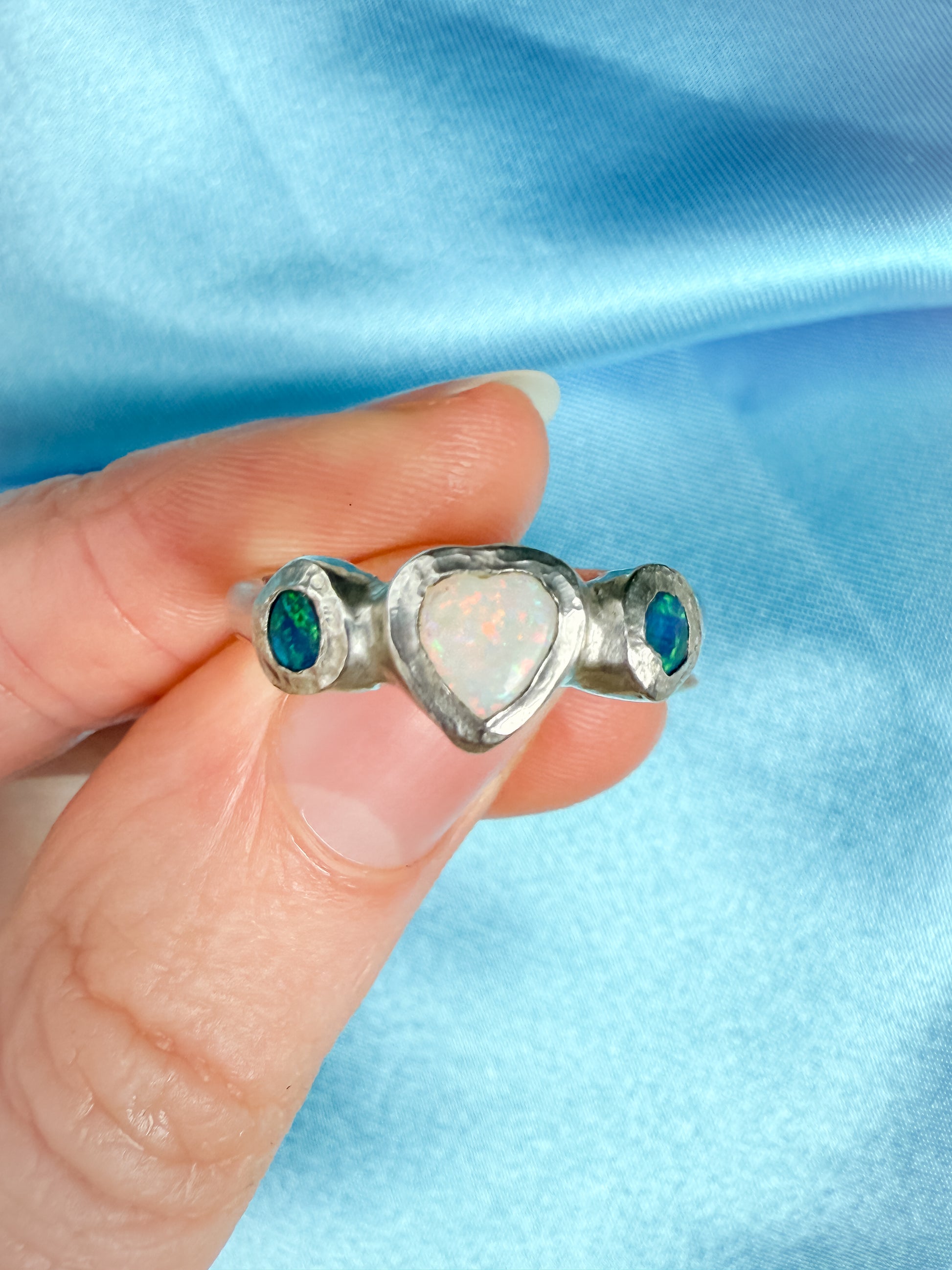 Silver ring with heart-shaped center and green gemstones held by a hand against a blue fabric background