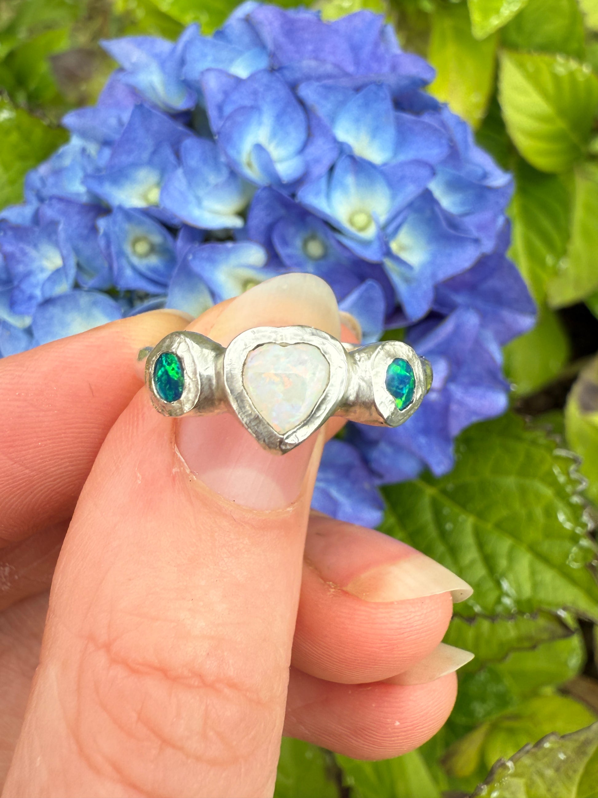 Silver heart ring with white and blue-green opals held against a background of blue flowers and green leaves