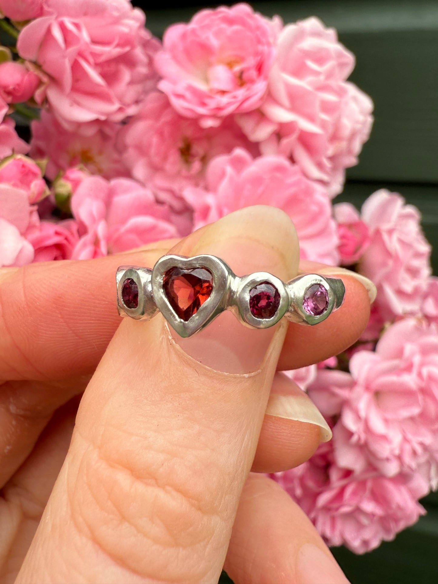 Silver ring with heart-shaped gemstones held against a background of pink flowers