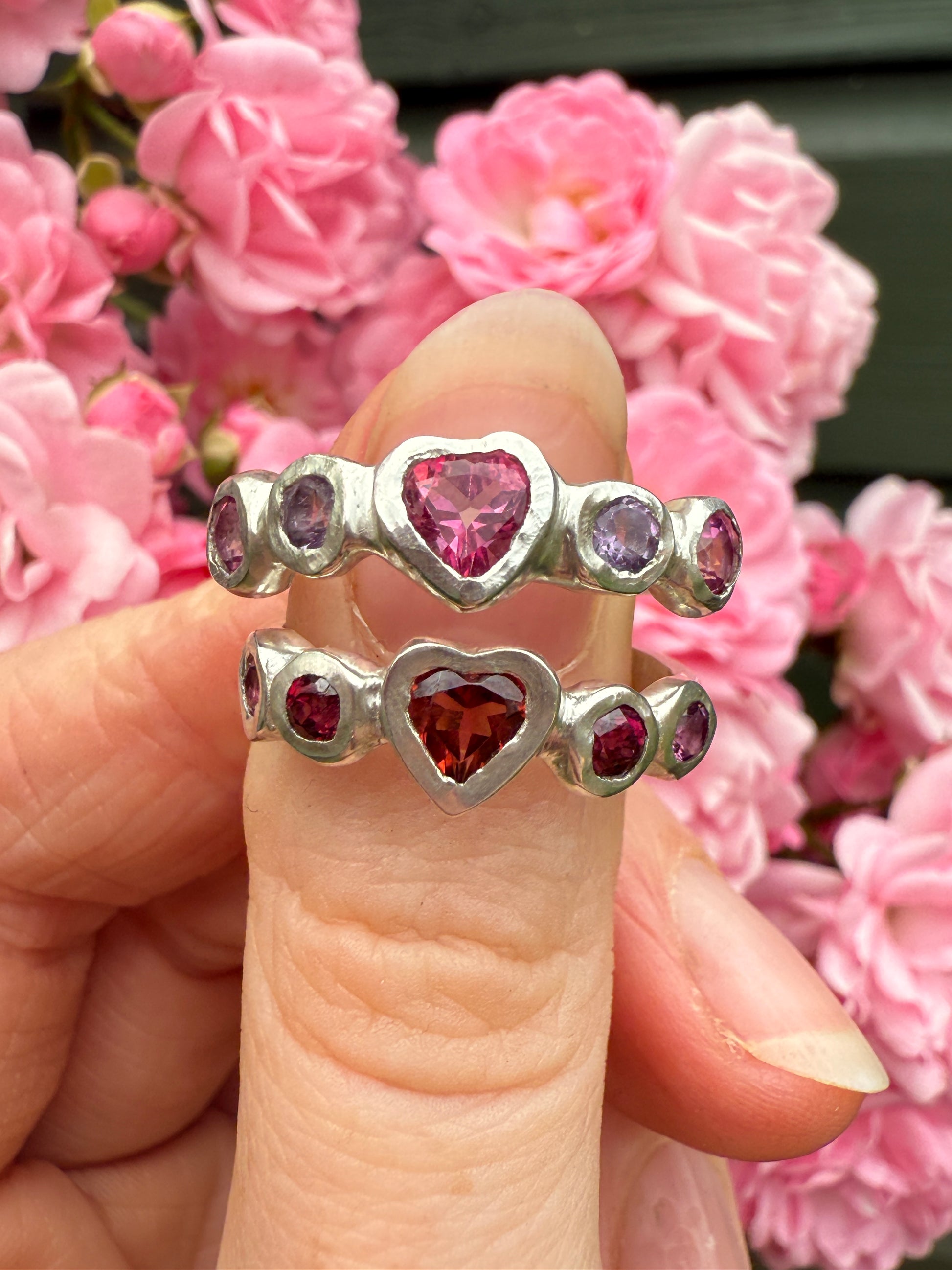 Silver rings with heart-shaped gemstones held between fingers against a pink floral background
