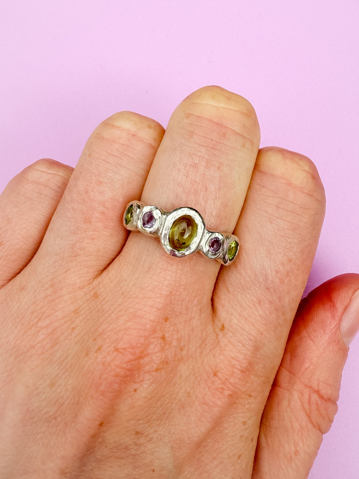 Silver bohemian ring with green and purple gemstones on a hand against a purple background