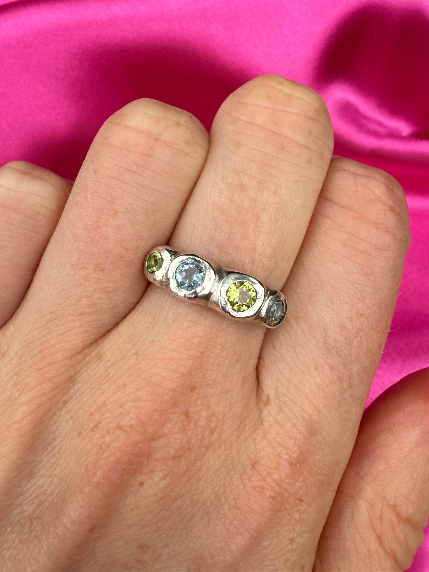 Silver bohemian ring with blue and green gemstones on a hand against a pink background
