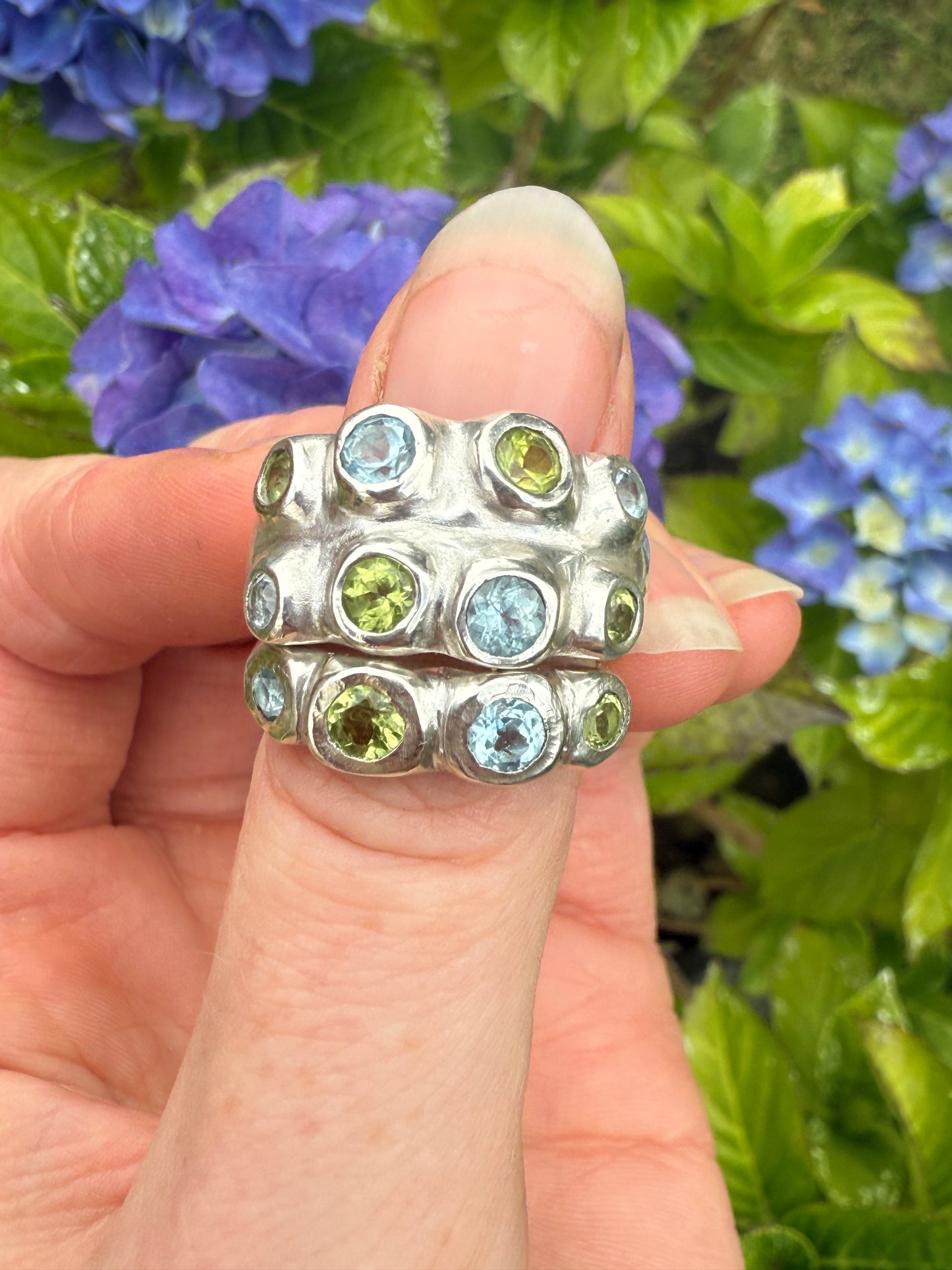 Silver rings with blue and green gemstones held against a background of blue flowers and green leaves.