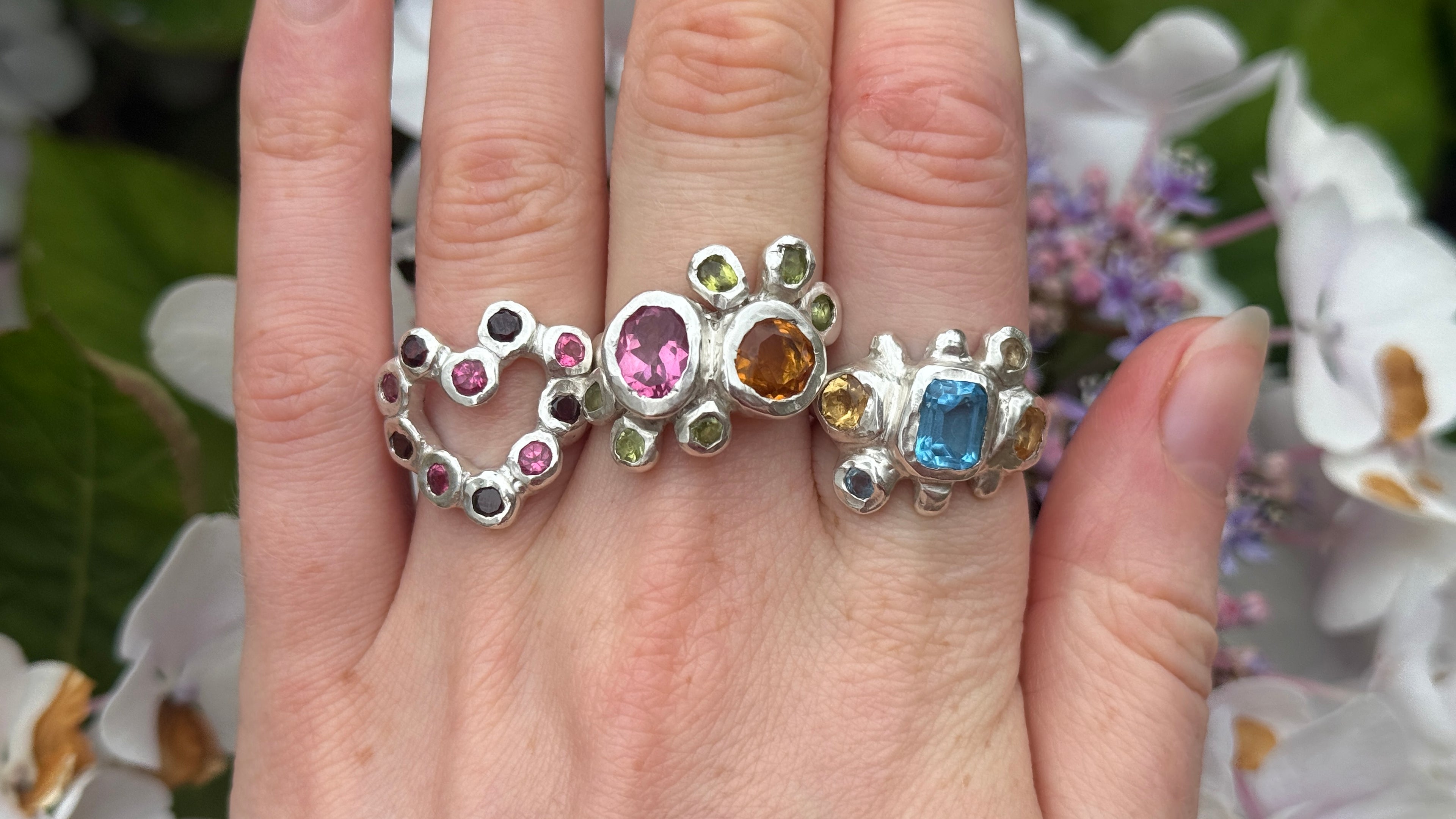 Three silver rings with colourful gemstones on a hand against a floral background