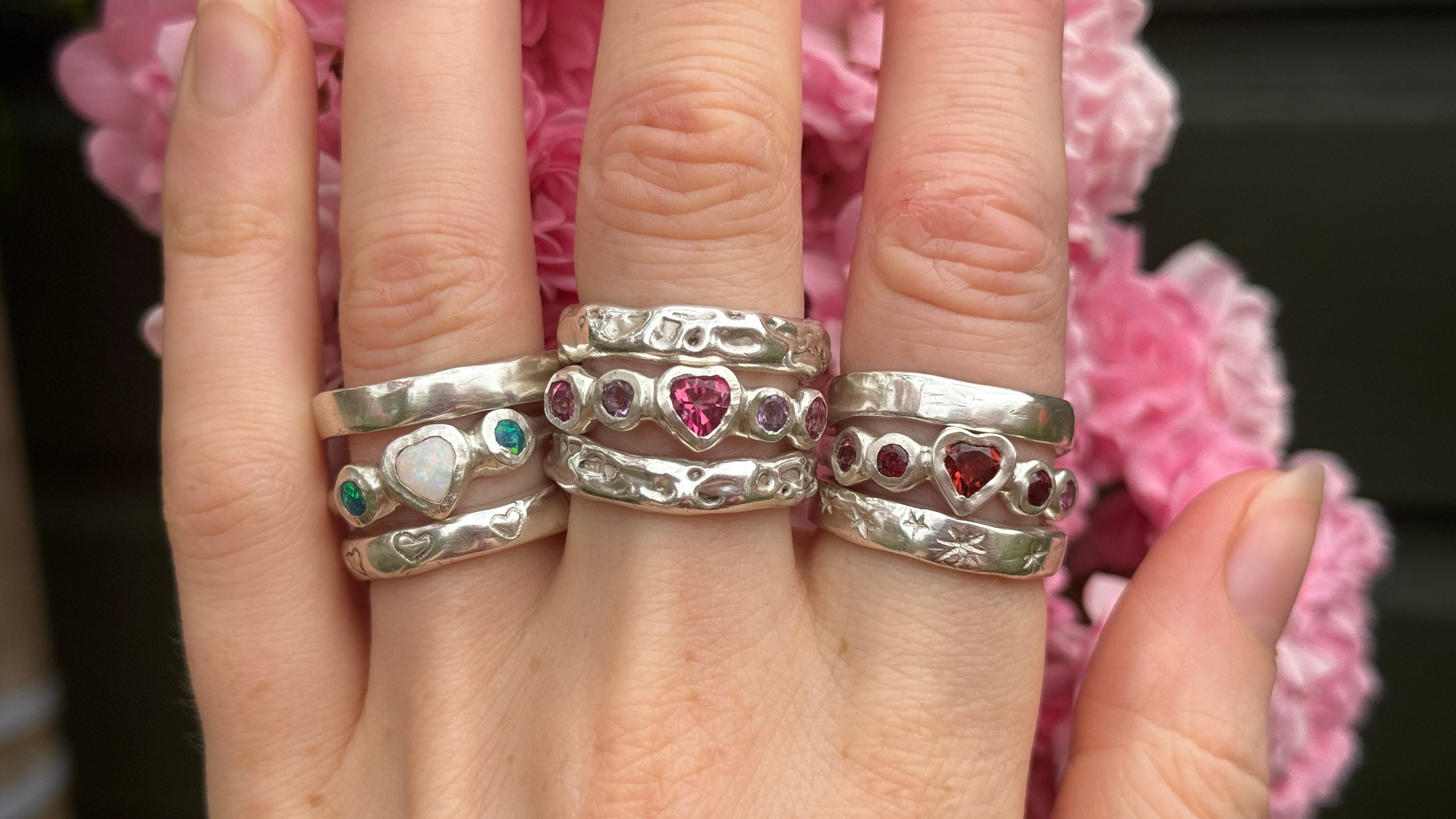 Hand wearing three silver rings with gemstones against a pink floral background