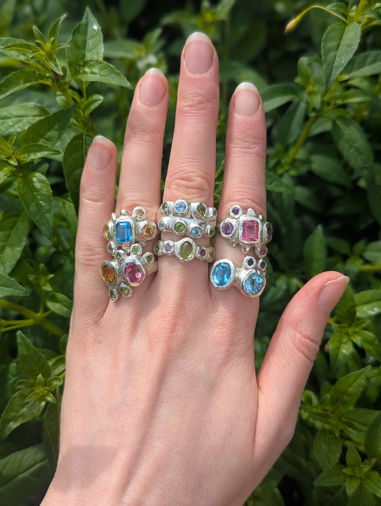 Hand wearing multiple rings with colorful gemstones against a green leafy background