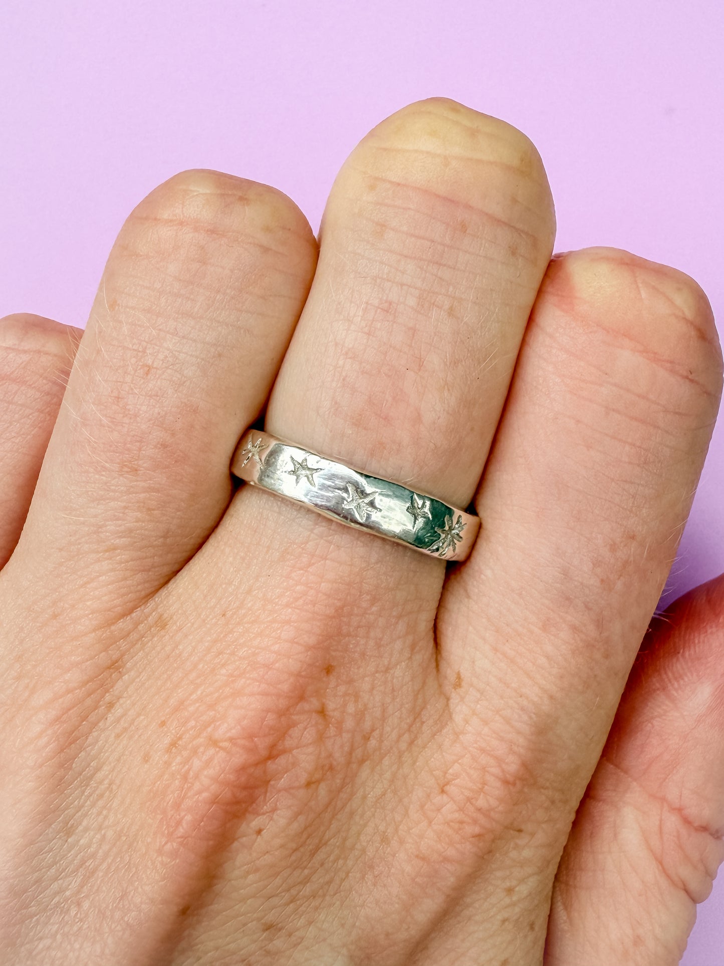 Silver ring with engraved star design on a finger against a pink background