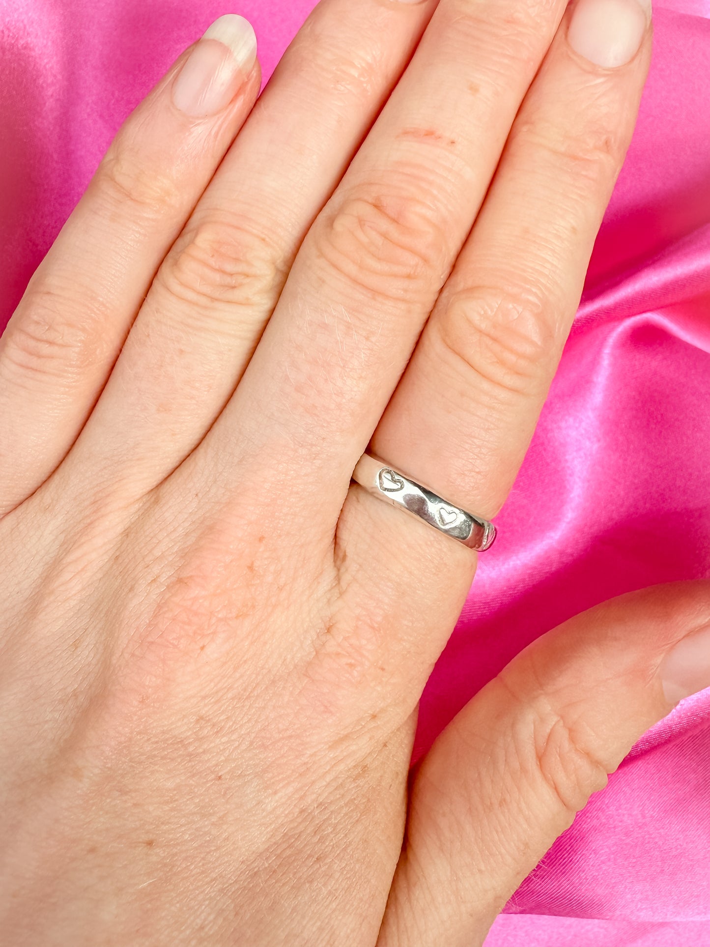 Hand wearing a silver ring with heart designs on a pink satin background