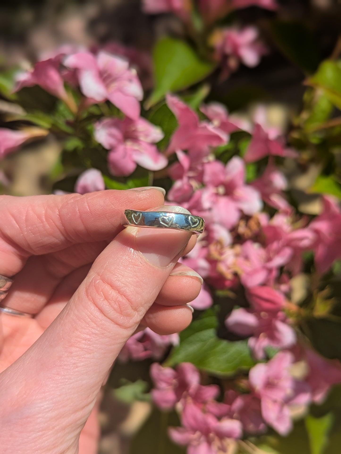 Hand holding a silver ring with etched hearts in front of pink flowers.