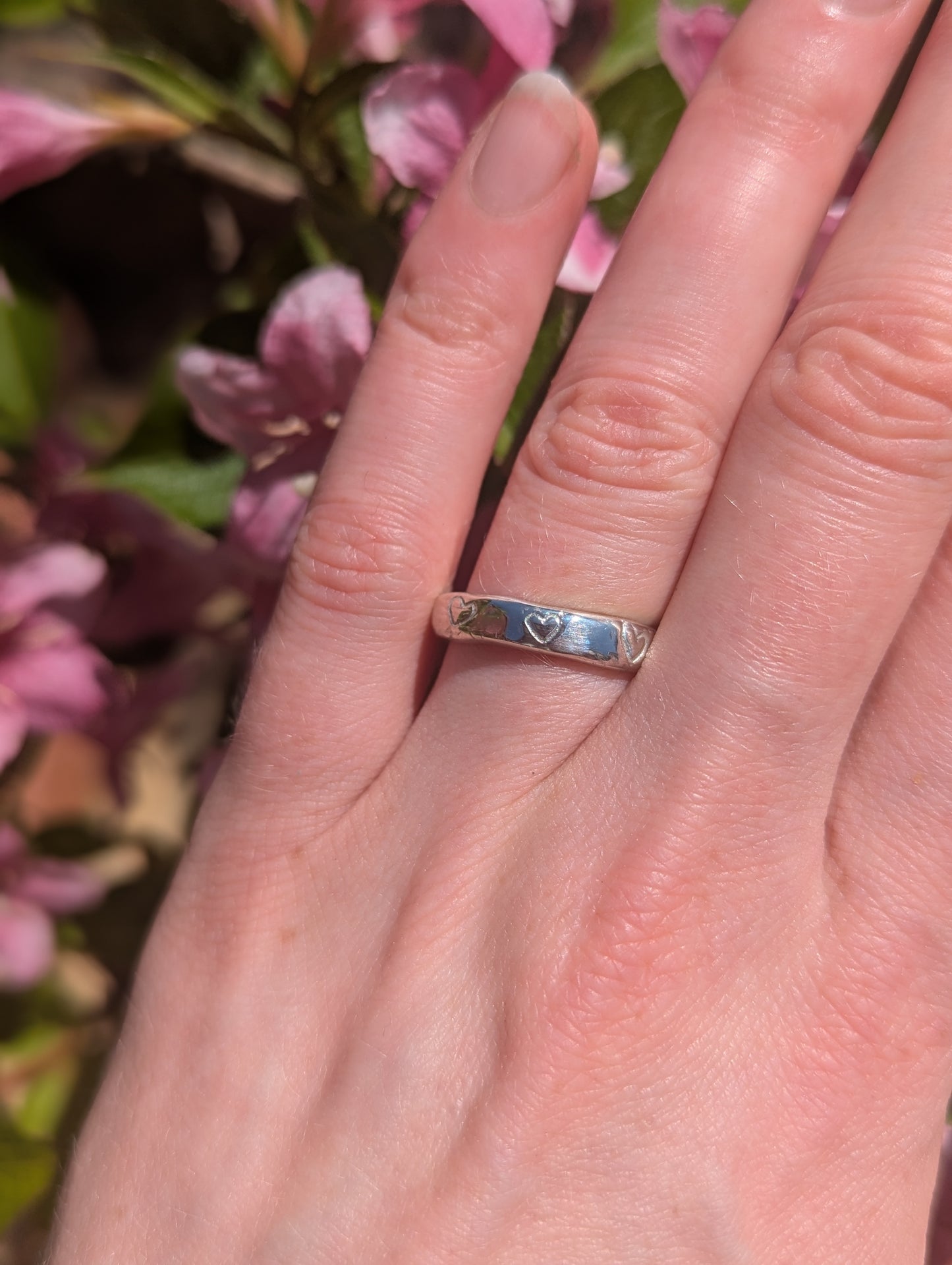 A silver stacking ring with etched hearts on a person's hand, with flowers in the background.