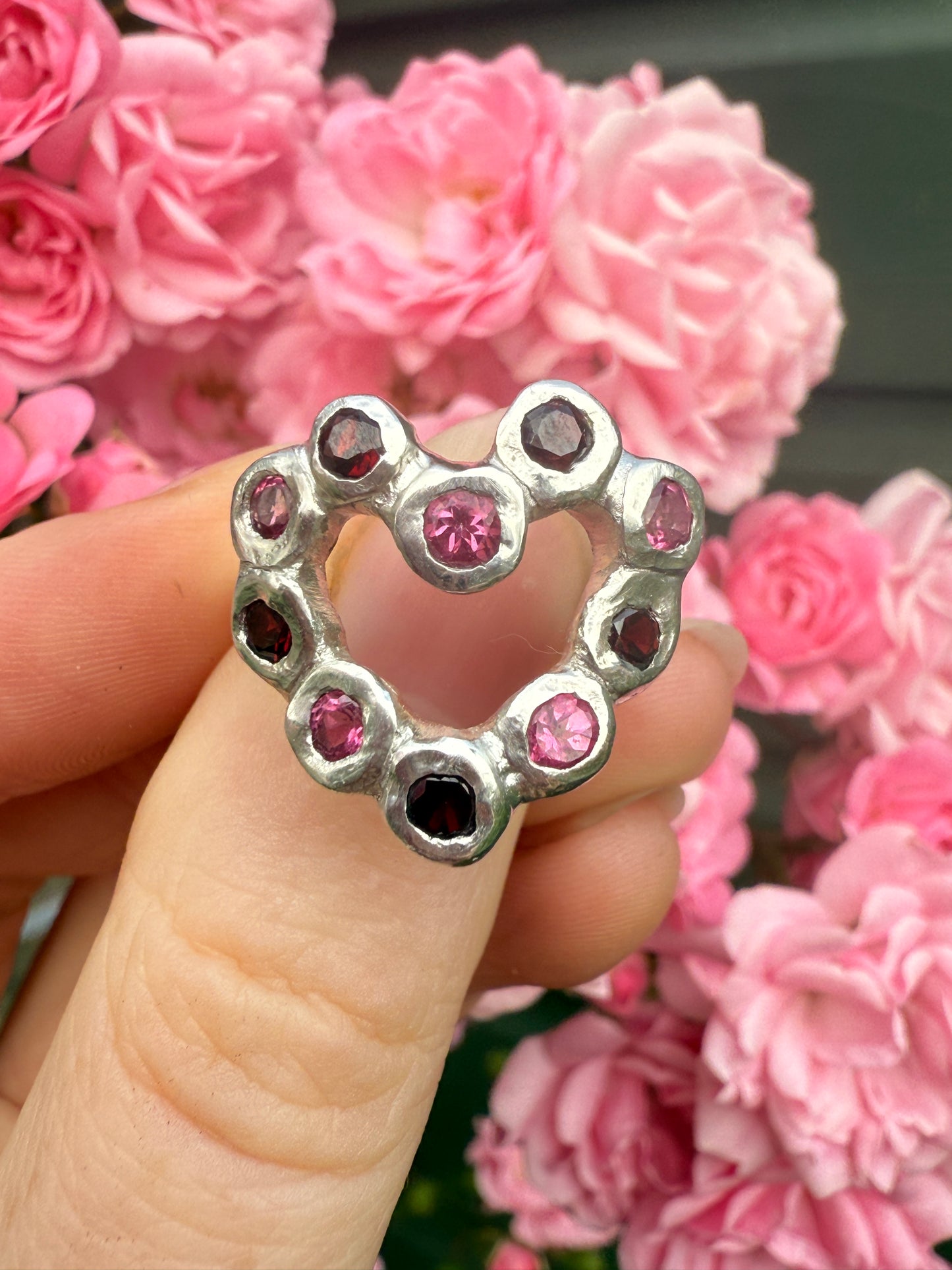 Silver heart ring with pink and red gemstones held in front of pink flowers