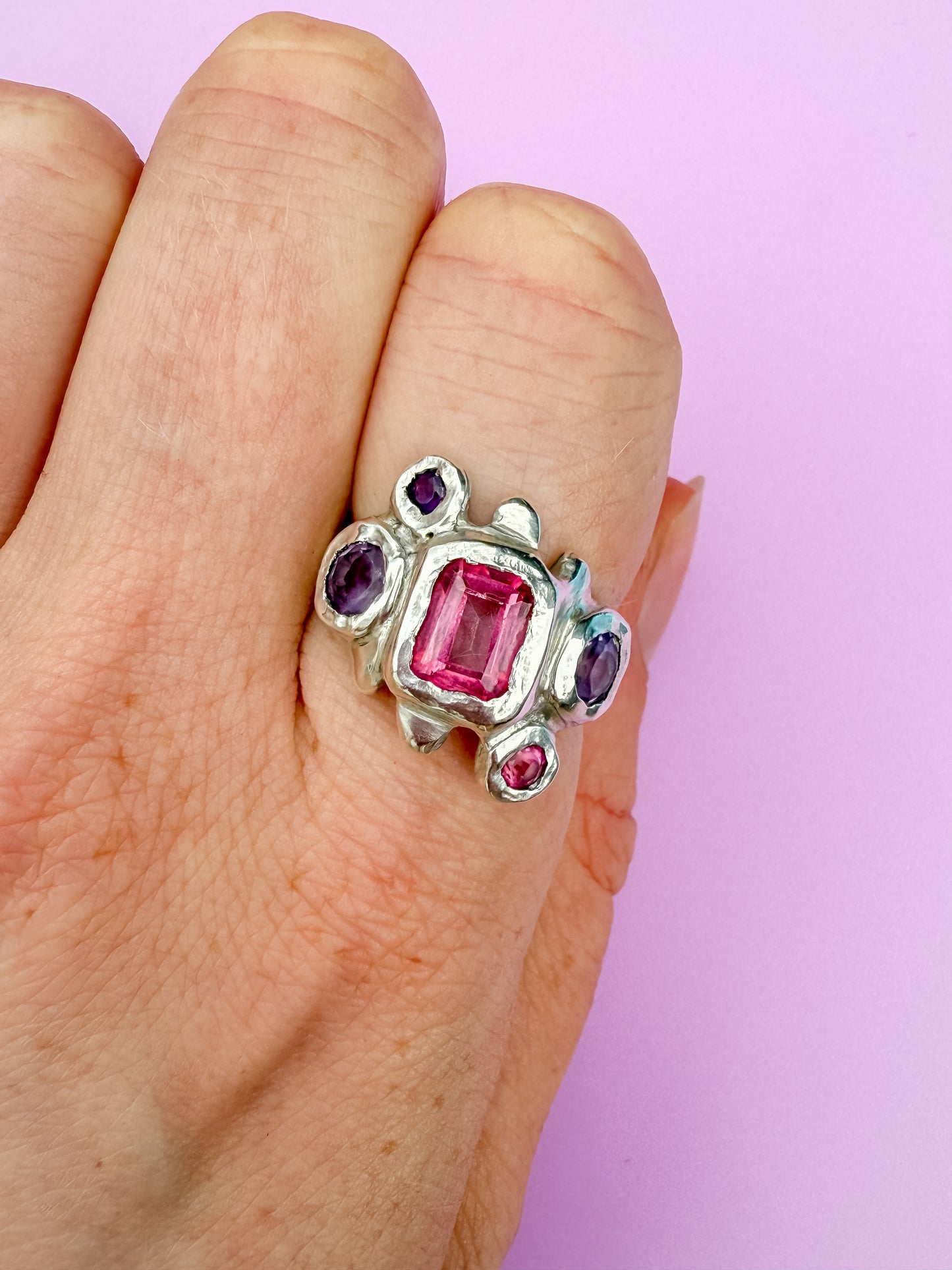 Silver Ring with pink and purple gemstones on a hand against a lilac background