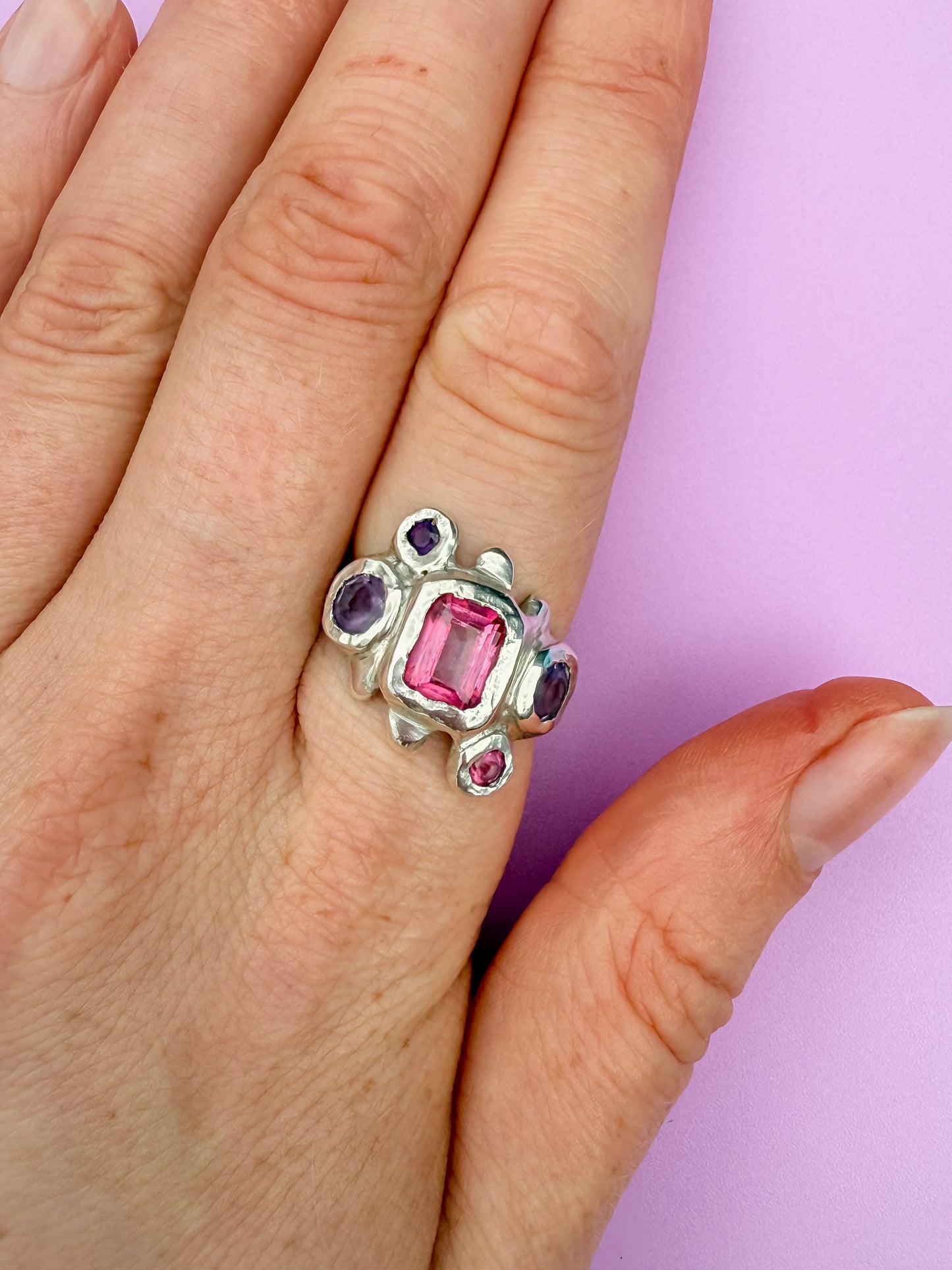 Silver bohemian ring with pink and purple gemstones on a hand against a light purple background