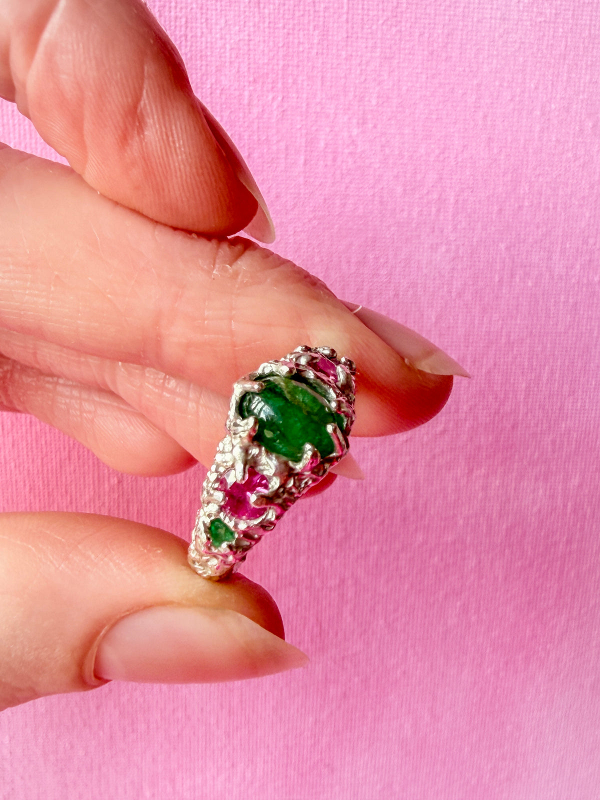 A close-up of a hand holding a silver ring with a large green emerald-cut center stone and small pink gemstone accents, set against a solid pink background.
