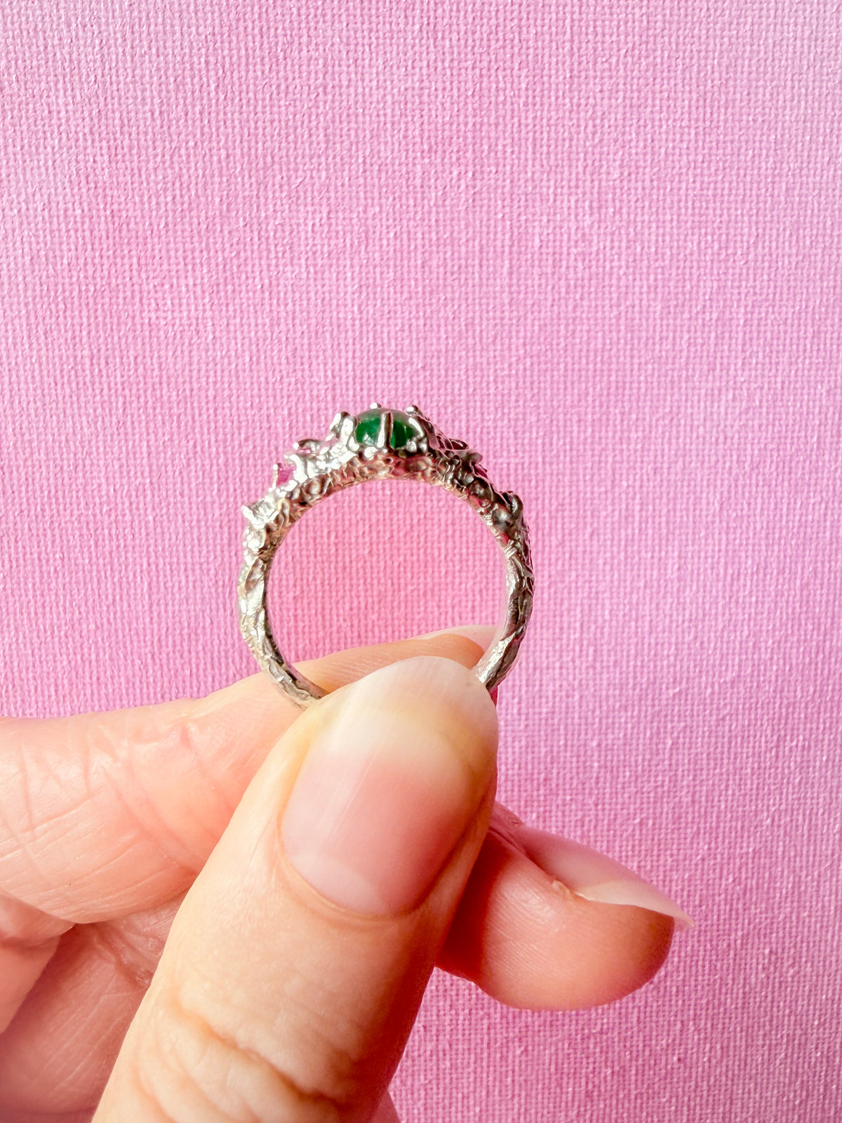 A silver-toned ring with a textured, organic band and a central cluster of green gemstones, held between fingers against a pink textured background.