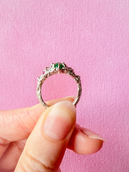 A silver-toned ring with a textured, organic band and a central cluster of green gemstones, held between fingers against a pink textured background.