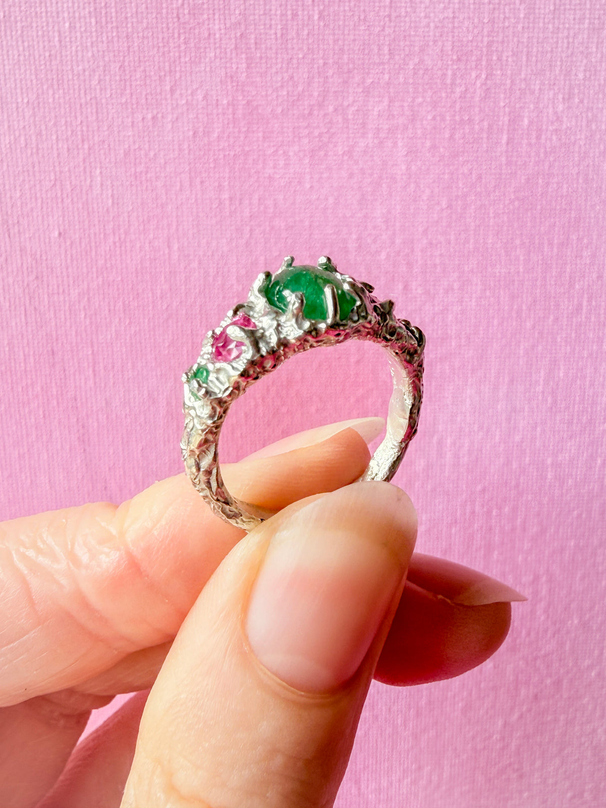 A hand holding a silver ring with a large green oval center stone and smaller pink and green accent stones, set against a solid pink background.
