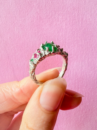 A hand holding a silver ring with a large green oval center stone and smaller pink and green accent stones, set against a solid pink background.