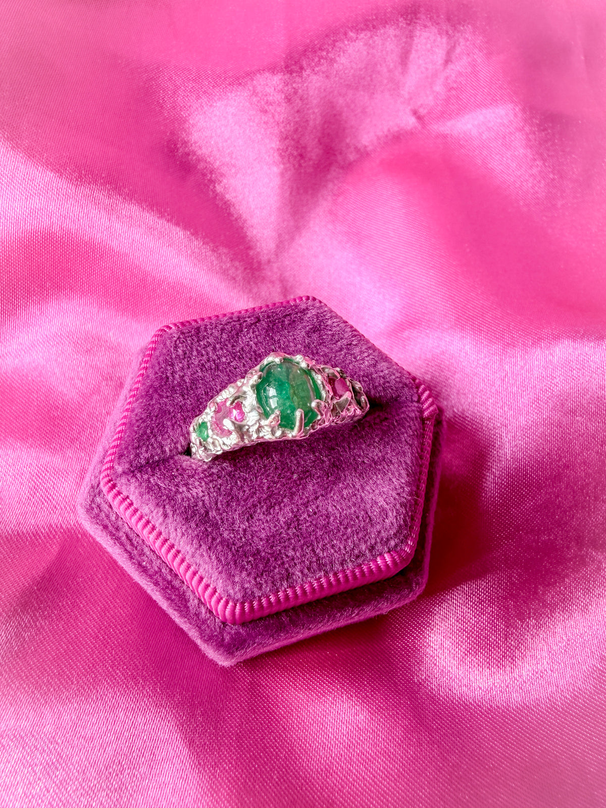A silver ring with a green emerald-cut center stone and pink gemstone accents, displayed on a hexagonal magenta velvet ring box over a pink satin fabric background.