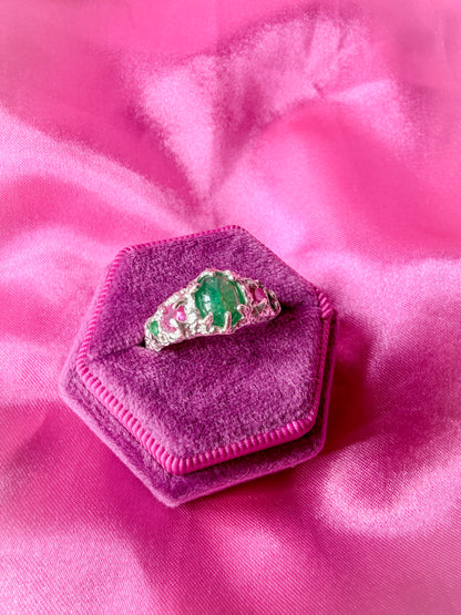A silver ring with a green emerald-cut center stone and pink gemstone accents, displayed on a hexagonal magenta velvet ring box over a pink satin fabric background.