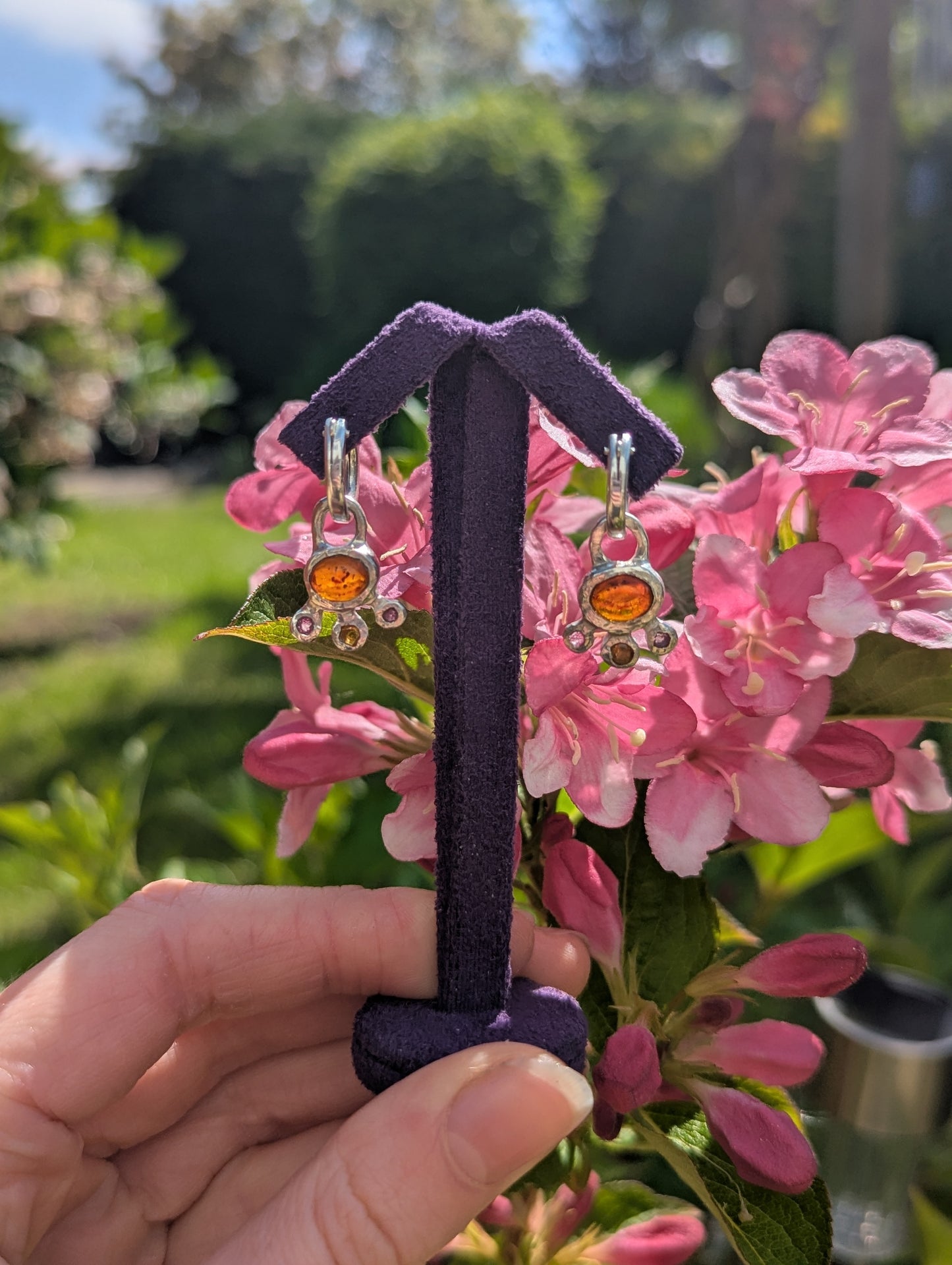 Pair of earrings on a purple stand held in front of pink flowers