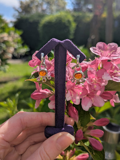 Pair of earrings on a purple stand held in front of pink flowers