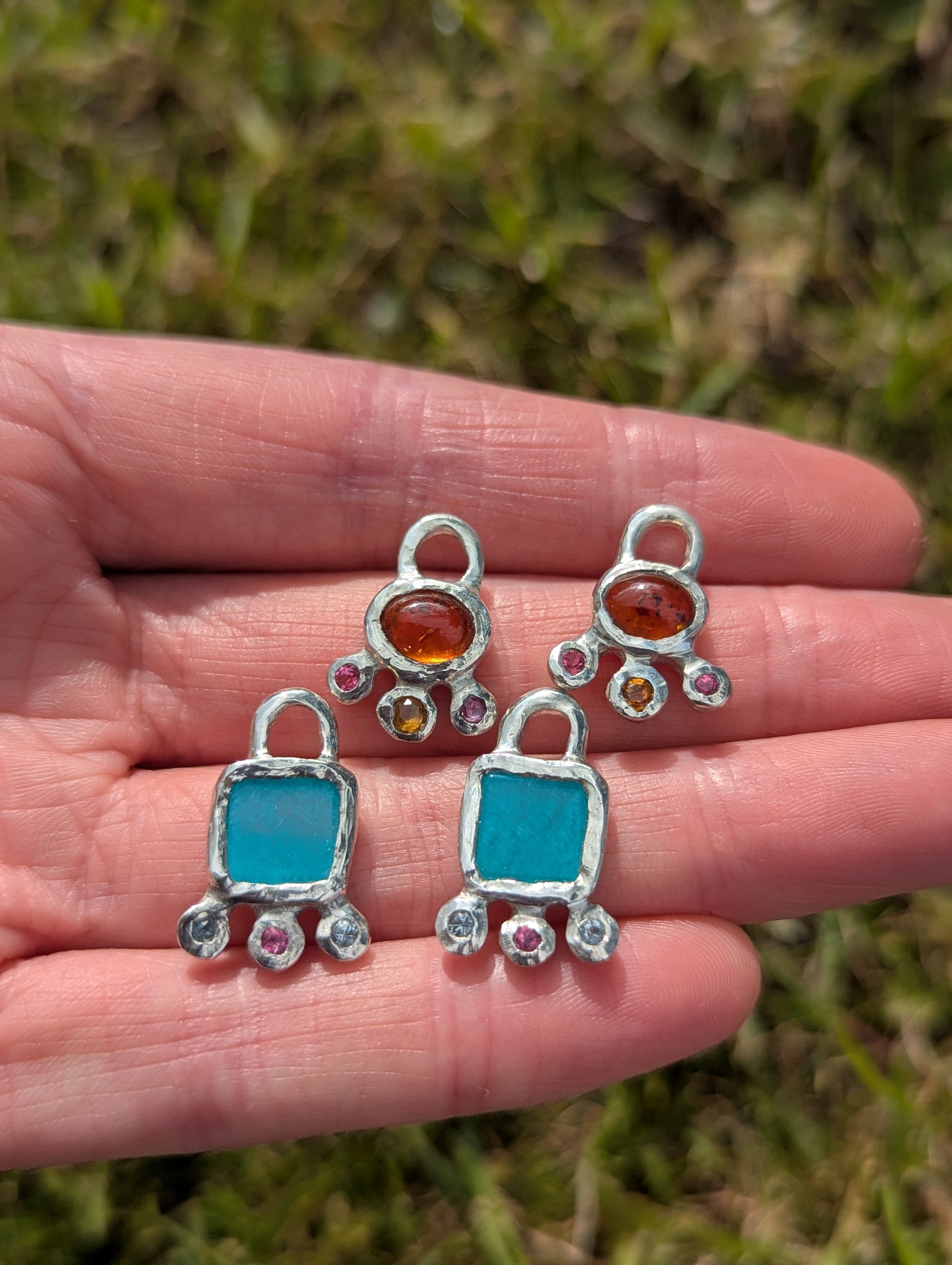 Four silver charms with blue stones and amber gemstones held in a hand against a grassy background.