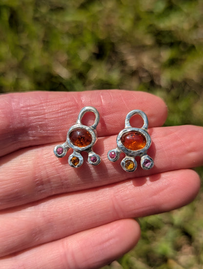 Two silver charms with colored stones held in a hand against a blurred green background