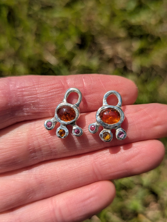 Two silver charms with colored stones held in a hand against a blurred green background