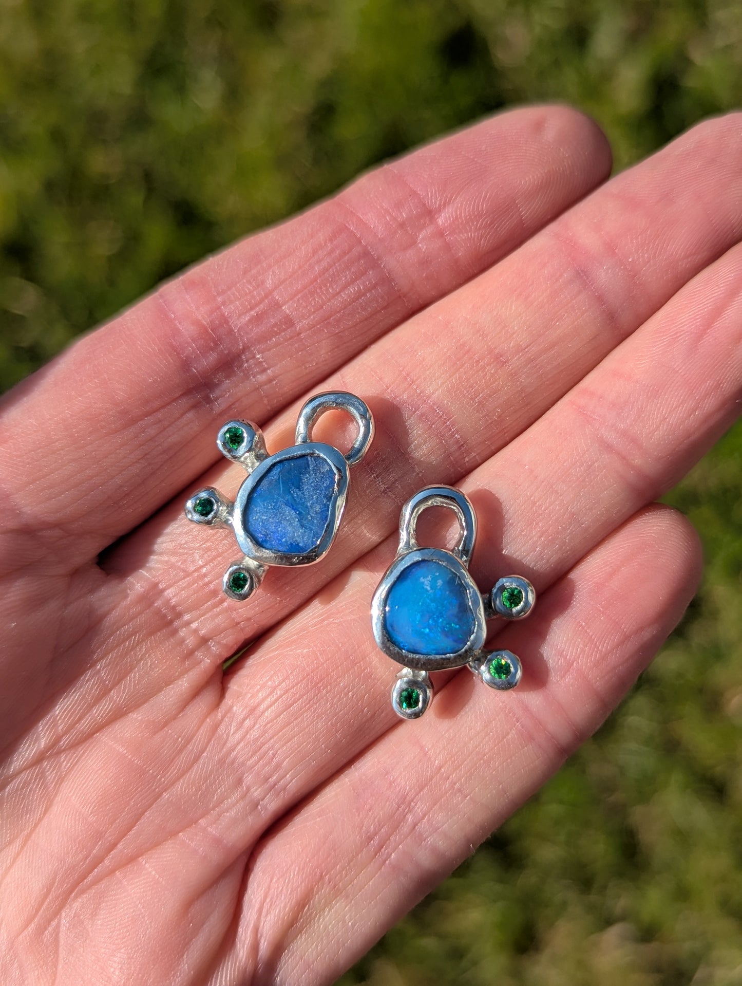 Two blue stone pendants with silver and green accents held in a hand against a blurred green background.