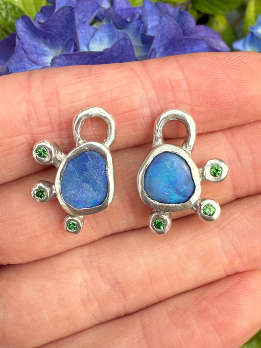 Two silver earring charms with blue stones and green gemstones on a hand next to purple flowers.