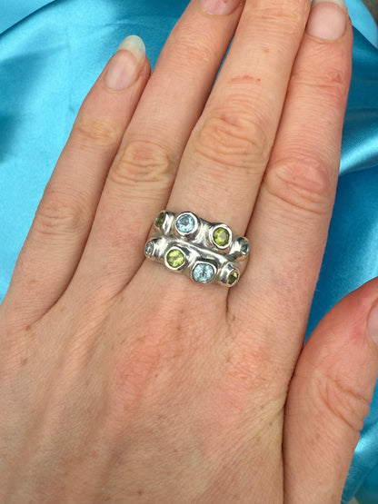 Silver bohemian ring with blue and green gemstones on a hand against a blue satin background