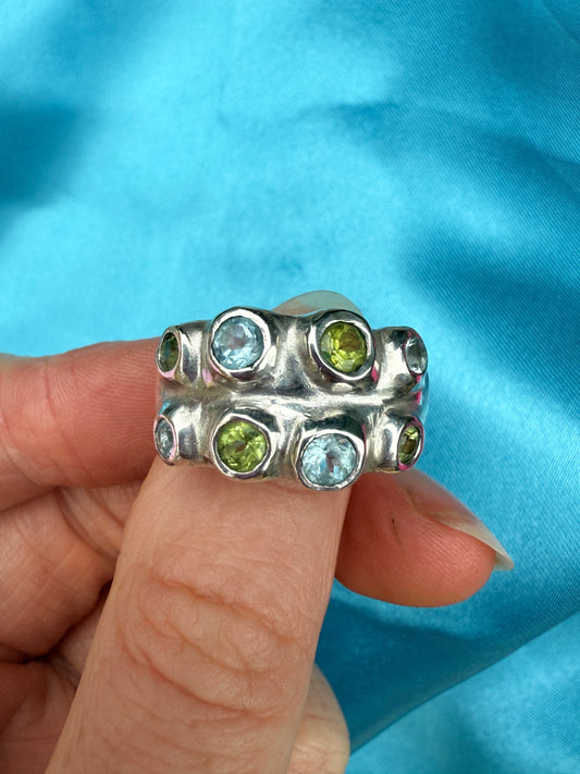 Silver bohemian ring with green and blue gemstones held by a hand against a blue fabric background