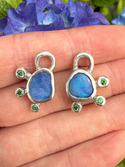 Two silver earring charms with blue stones and green gemstones on a hand next to purple flowers.