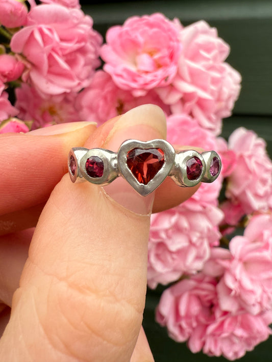 Silver heart ring with red and pink heart gemstones held between fingers against pink flowers