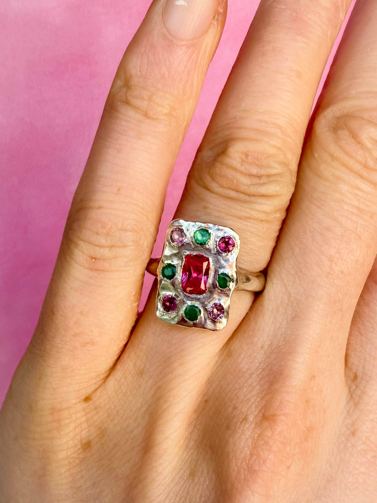 Oblong silver handmade Ring with red, green and pink gemstones on a hand against a pink background