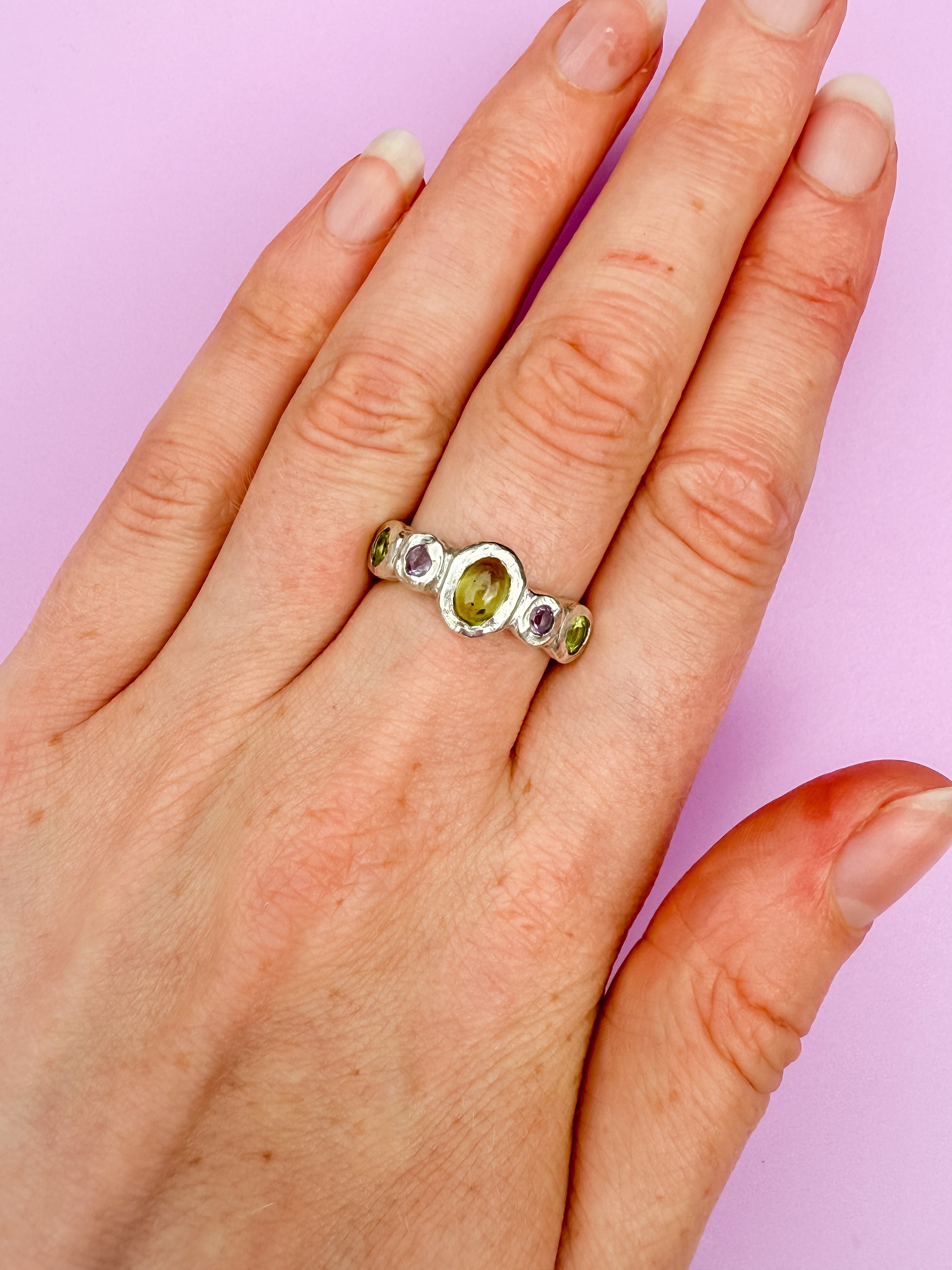 Silver bohemian ring with green and purple gemstones on a hand against a purple background