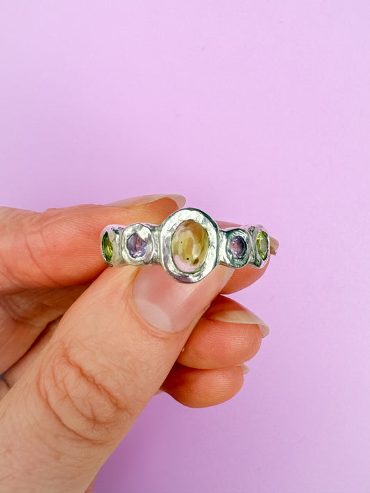 Silver bohemian ring with green and purple gemstones on a hand against a purple background
