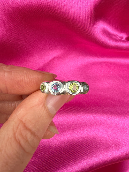 Silver bohemian ring with blue and green gemstones held between fingers against a pink fabric background