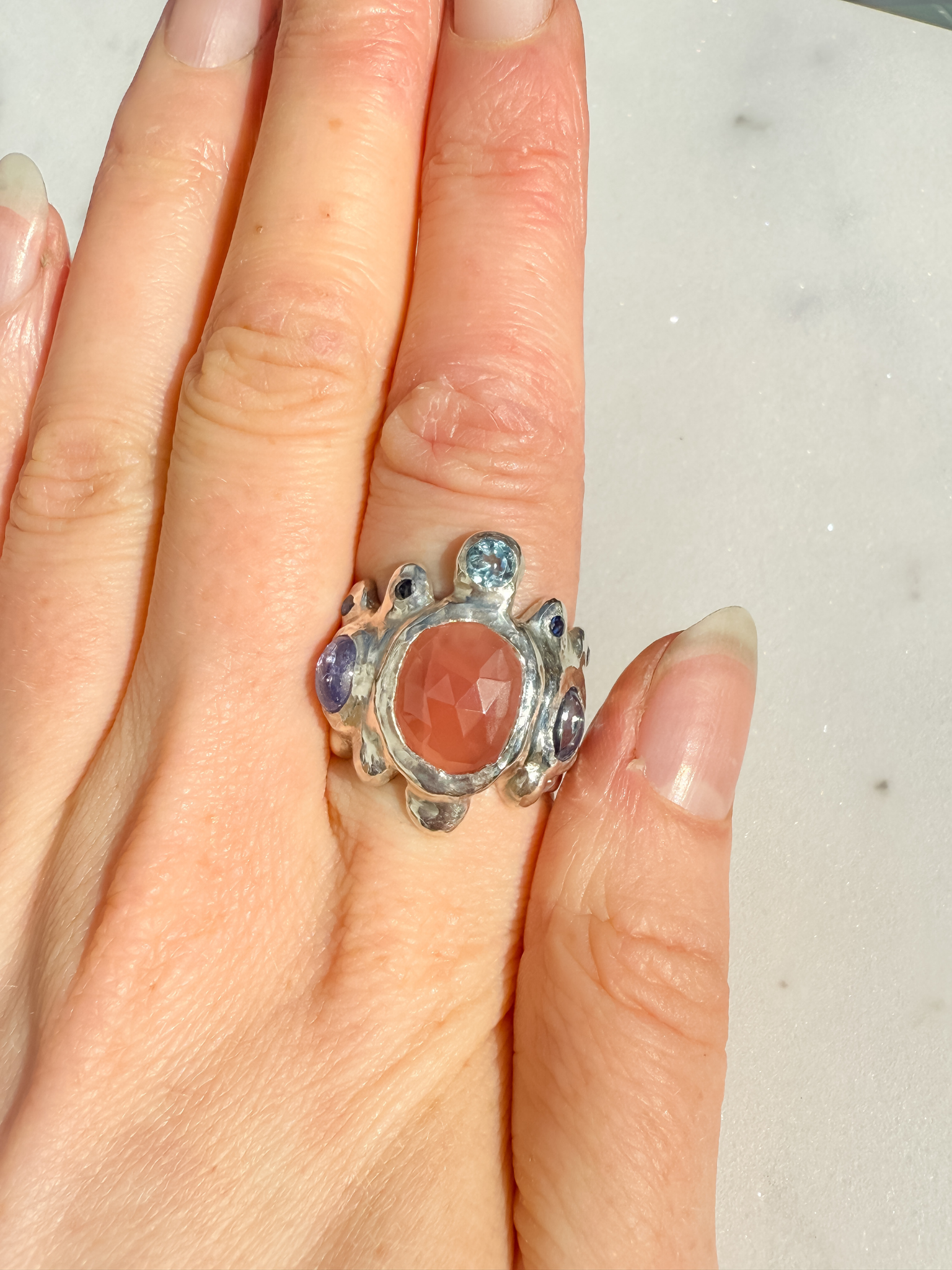 Silver ring with a large central stone and smaller stones on a finger against a neutral background