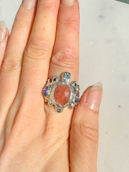 Silver ring with a large central stone and smaller stones on a finger against a neutral background