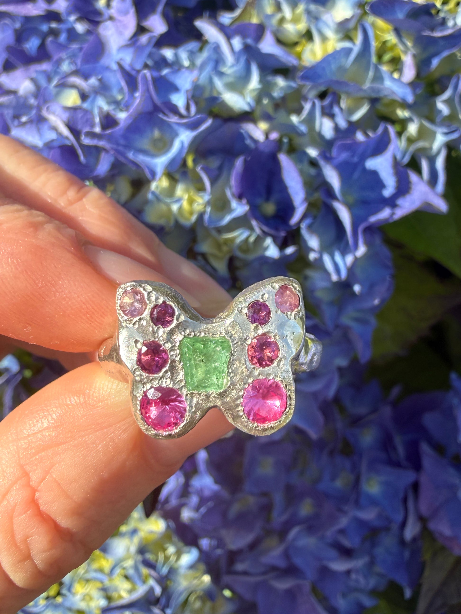 Butterfly-shaped ring with colorful stones held in front of blue flowers