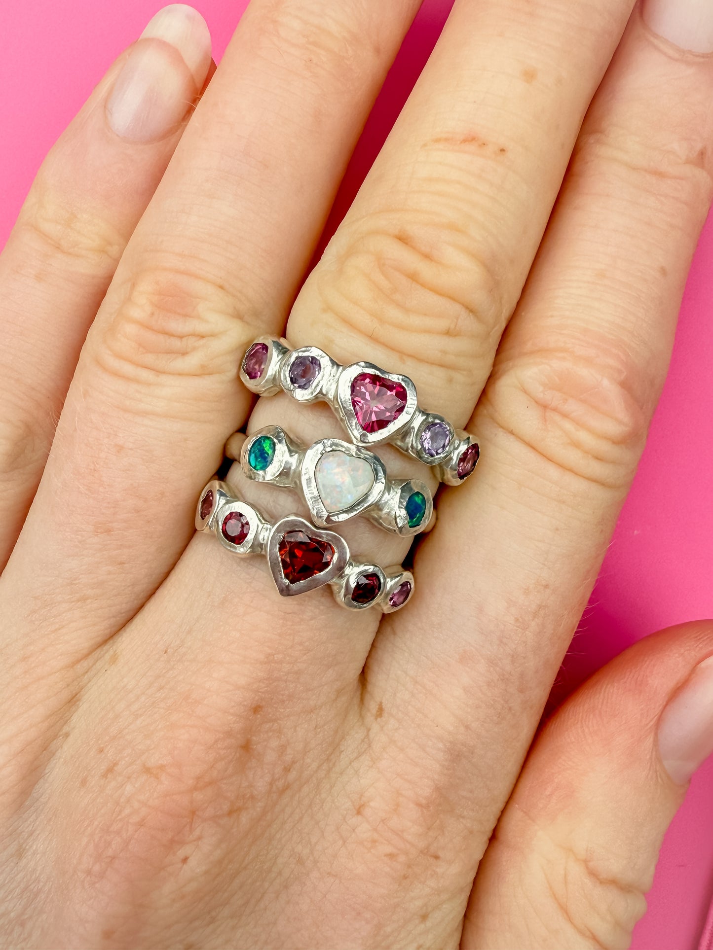 Three silver rings with heart shaped gemstones in pink, white, red, and purple