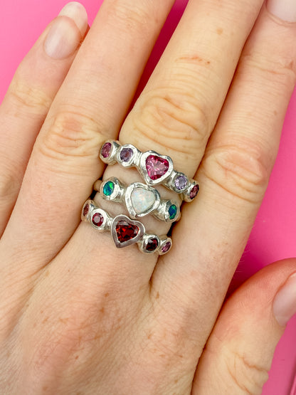 Three silver rings with heart shaped gemstones in pink, white, red, and purple