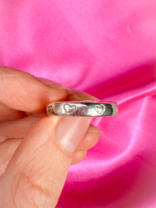 Silver ring with heart designs held by a hand against a pink fabric background